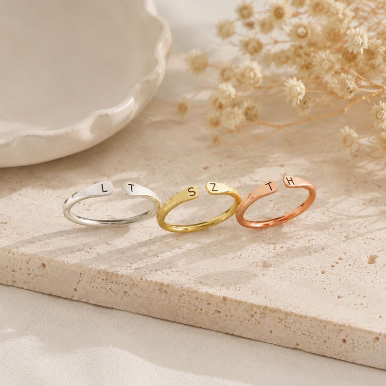 May include: Three open-ended rings in silver, gold, and rose gold. Each ring features a single initial: L, S, and T on the silver ring; S and Z on the gold ring; and T and H on the rose gold ring. The rings are displayed on a neutral-toned surface.