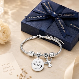 Peut inclure: Un bracelet en cha&icirc;ne serpent argent&eacute;e avec deux perles cylindriques grav&eacute;es "LOVE" et un c&oelig;ur, un pendentif rond grav&eacute; "Tracy" et "1940-2026" avec des ailes d'ange au-dessus, et une breloque ange, pr&eacute;sent&eacute; avec une bo&icirc;te cadeau bleu marine.