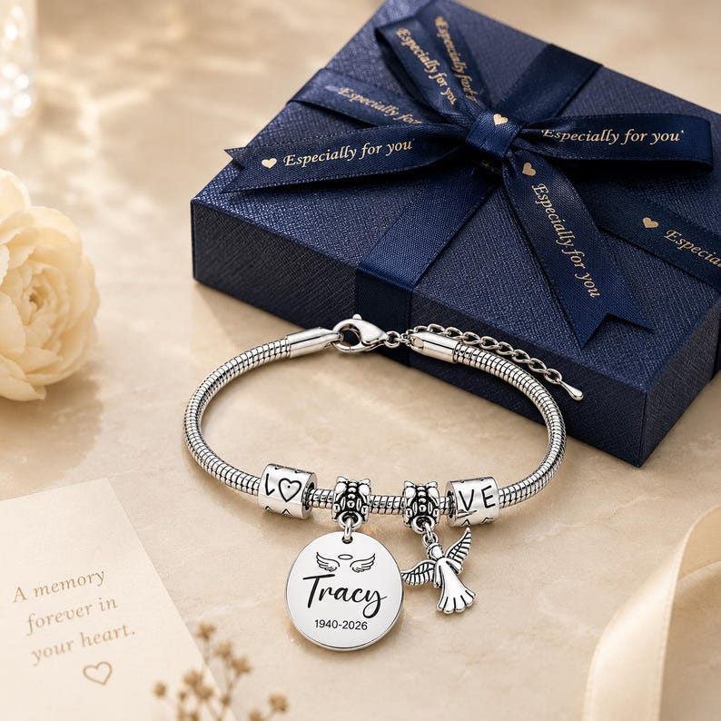 Peut inclure: Un bracelet en cha&icirc;ne serpent argent&eacute;e avec deux perles cylindriques grav&eacute;es "LOVE" et un c&oelig;ur, un pendentif rond grav&eacute; "Tracy" et "1940-2026" avec des ailes d'ange au-dessus, et une breloque ange, pr&eacute;sent&eacute; avec une bo&icirc;te cadeau bleu marine.