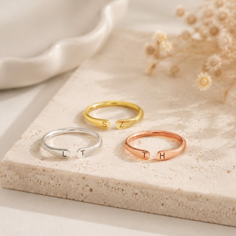 May include: Three open-ended rings in silver, gold, and rose gold. Each ring has a single letter engraved on each end. The rings are displayed on a neutral-toned surface with dried flowers in the background.