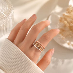 May include: A multi-tone ring with the letters L, T, S, and H engraved on the bands. The ring has a silver, gold, and rose gold finish. The ring is worn on a finger.