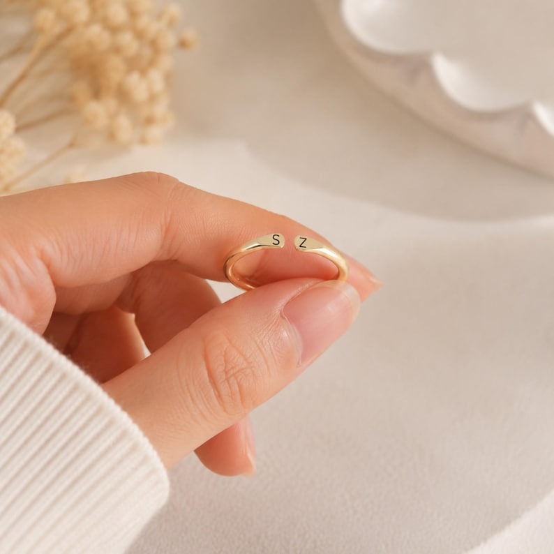 May include: A gold-colored ring with the letters "S" and "Z" engraved on the top. The ring is held between fingers, showcasing its simple, elegant design. The ring is a delicate piece of jewelry.