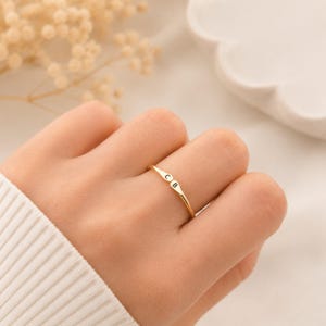 May include: A gold-colored ring with the initials "C B" engraved on a rectangular plate. The ring is on a finger, and the background is blurred, with a white sweater sleeve visible.
