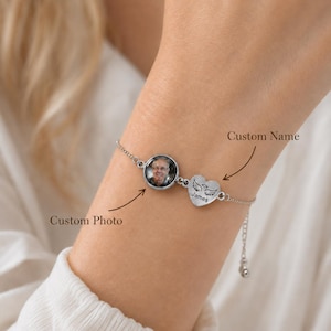 Puede incluir: Una pulsera de cadena en tono plateado con un dije circular con una foto personalizada de una persona y un dije en forma de corazón grabado con el nombre James y un par de alas de ángel. La pulsera incluye etiquetas de texto indicativas.