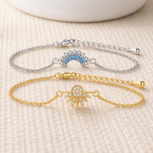 May include: Two delicate bracelets with fine chains. The top silver-tone bracelet features a crescent moon charm set with clear and light blue stones. The bottom gold-tone bracelet features a sunburst charm set with clear stones and a lobster clasp.