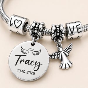 Peut inclure: Un bracelet &agrave; breloques argent&eacute; avec un pendentif rond grav&eacute; du nom Tracy, des ann&eacute;es 1940-2026, et une paire d'ailes d'ange avec une aur&eacute;ole. Le bracelet comprend une petite breloque ange et deux perles cylindriques portant l'inscription LOVE.