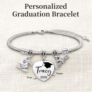 May include: A silver-toned graduation bracelet with charms. The bracelet features a graduation cap charm, a diploma charm, and a heart-shaped charm engraved with "Tracy" and "2026". The text "Personalized Graduation Bracelet" is at the top.