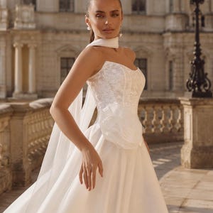 May include: A strapless white wedding dress featuring a lace bodice with a sweetheart neckline, a large fabric flower accent at the waist, and a full-length skirt made of layered tulle. A matching sheer white scarf is draped around the neck.