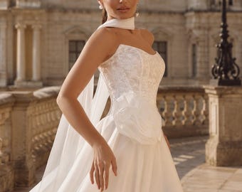 May include: A strapless white wedding dress featuring a lace bodice with a sweetheart neckline, a large fabric flower accent at the waist, and a full-length skirt made of layered tulle. A matching sheer white scarf is draped around the neck.