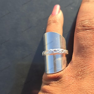 Pode incluir: Um anel prateado com um design único. O anel apresenta uma faixa larga e retangular que cobre parte do dedo. Uma faixa menor com um padrão floral também é visível. O anel é usado em um dedo.