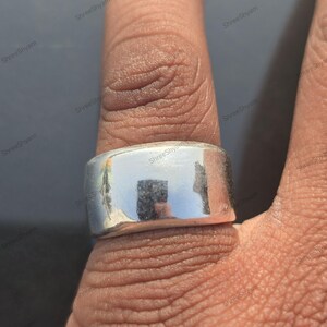 Op de afbeelding: Een zilverkleurige ring met een brede band wordt op een vinger gedragen. De ring heeft een glad, reflecterend oppervlak. De ring is een eenvoudig, modern ontwerp, geschikt voor dagelijks gebruik. De ring is waarschijnlijk van metaal.