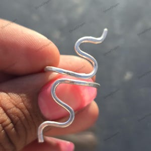 Op de afbeelding: Een zilverkleurige ring in de vorm van een slang. De ring heeft een kronkelend, serpentijnachtig ontwerp dat zich om de vinger wikkelt. De ring wordt tegen een vage achtergrond gehouden.