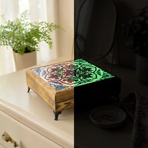 May include: Wooden jewelry box with a colorful, patterned tile top. The box has small black feet and a light wood finish. The top glows green in the dark. The box is on a white surface.