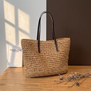 May include: A woven straw tote bag with dark brown leather handles. The bag is rectangular with a wide opening. The text "Metaphorical Concepts" is visible on a book in the background. The bag is sitting on a wooden surface.