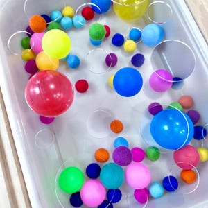 May include: A collection of colorful balls and clear circular shapes in a white container. The balls are various sizes and colors, including red, blue, green, yellow, pink, and orange. Some balls are smooth, while others have a fuzzy texture.