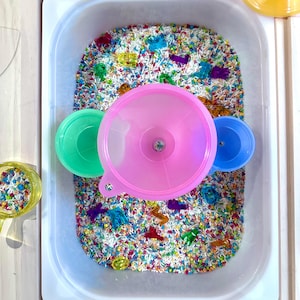 K&ouml;nnte beinhalten: Eine sensorische Wanne gef&uuml;llt mit buntem Reis und kleinen Plastikspielzeugen. Ein rosa Trichter, ein gr&uuml;ner Deckel und ein blauer Deckel liegen auf dem Reis. Die wei&szlig;e, rechteckige Wanne ist ideal f&uuml;r Kinderspiele.