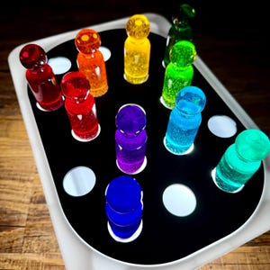 May include: A board game with a black surface and a white border. The game features colorful, translucent game pieces in shades of red, orange, yellow, green, blue, and purple. The pieces are arranged on the board with circular cutouts.
