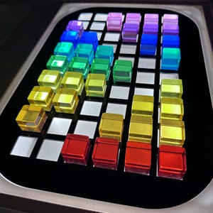 May include: A close-up of a black surface with a grid of clear, colorful, square blocks. The blocks are arranged in rows and columns, with colors ranging from red to purple. The blocks are made of a translucent material and appear to be illuminated.
