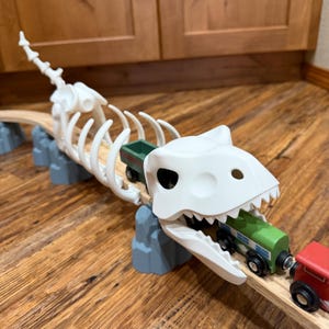 May include: A white dinosaur skeleton toy bridge with a wooden train track. The dinosaur's mouth holds a green train car, and a red train car is visible. The bridge is supported by gray rock-shaped structures.