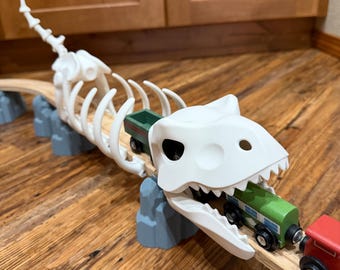 Può includere: Un ponte giocattolo a forma di scheletro di dinosauro bianco con un binario di legno. La bocca del dinosauro tiene un vagone verde, e un vagone rosso è visibile. Il ponte è sostenuto da strutture grigie a forma di roccia.