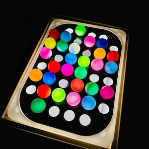 May include: A black tray with a gold border, filled with an array of colorful, spherical objects. The spheres are in various bright colors, including red, pink, green, blue, and yellow. White circular accents are also present.