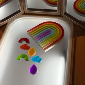 May include: A colorful wooden rainbow arch toy with multiple layers of red, orange, yellow, green, blue, and purple. Several small wooden cloud and raindrop shapes are scattered around the rainbow. The toy is on a white surface.