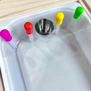 May include: A sensory play set featuring a white plastic bin with a clear divider, a small stainless steel cup, and four colorful droppers in pink, red, yellow, and green. The set is designed for early childhood development and learning.