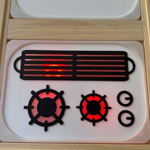 May include: A white stovetop play set featuring a black rectangular grill grate and two circular burner grates with bright red LED lighting underneath. Two small black circular control knobs are positioned to the right of the burners.