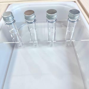 May include: Four clear glass test tubes with silver caps are arranged in a row. The tubes are held in place by a clear acrylic holder, set inside a white tray. The caps are silver and the tubes are empty.