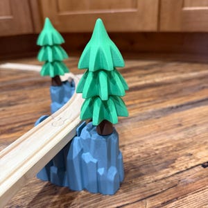 May include: Wooden toy train track bridge with two green tree decorations on blue rock formations. The track is light brown, and the trees are a vibrant green. The bridge is designed for toy trains.