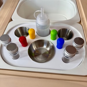 May include: A white sensory play tray with a variety of items. Includes a clear plastic bottle, stainless steel bowls, and colorful silicone bottle stoppers in red, yellow, green, and blue. Also includes small metal containers with silver lids.