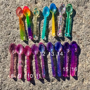 May include: Sixteen colorful resin spoons in various shades of pink, purple, blue, green, yellow, and orange. Each spoon has a unique design, some with glitter or embedded details. The spoons are numbered 1 through 16.