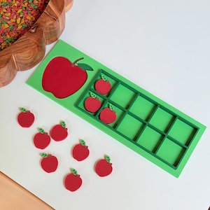May include: A bright green ten-frame counting tray with a large red apple cutout on the left side. The tray includes ten rectangular compartments, three of which contain small red apple tokens. Seven additional red apple tokens lie nearby.