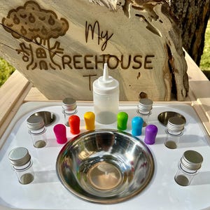May include: A sensory play station with a wooden sign that reads "My Treehouse". The station includes a stainless steel bowl, clear bottles with silver lids, and colorful bottle toppers in red, yellow, green, blue, and purple.