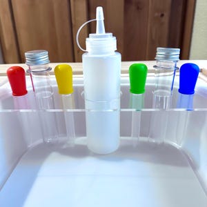 May include: A collection of clear glass test tubes with colorful stoppers in red, yellow, green, and blue. A white plastic bottle with a dispensing cap is centered, and the items are arranged in a white plastic tray.