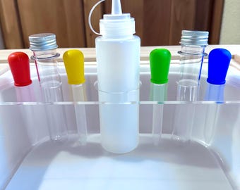 Könnte beinhalten: Eine Sammlung klarer Reagenzgläser mit farbigen Stöpseln in Rot, Gelb, Grün und Blau. Eine weiße Plastikflasche mit Dosierkappe ist mittig angeordnet, alles in einer weißen Kunststoffschale.