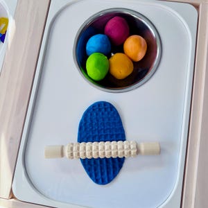 May include: A white play table with a stainless steel bowl containing colorful play dough. A blue textured mat and a wooden roller are also on the table. The play dough is in shades of blue, green, pink, orange, and yellow.