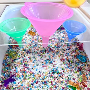 May include: A sensory bin with colorful rice and various small toys. Three plastic funnels in pink, green, and blue are positioned above the rice. The bin is designed for children's play and exploration, promoting tactile and visual stimulation.