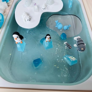May include: A sensory play tray with a light blue liquid base, featuring miniature arctic animal figurines. The tray includes penguins, polar bears, whales, and ice-like shapes, creating a winter wonderland theme. The tray is set in a light-colored wooden frame.