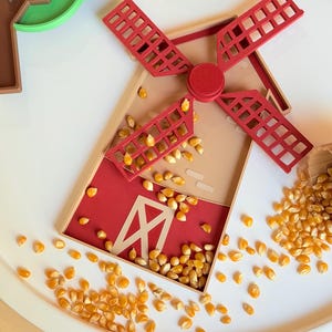 May include: A 3D-printed windmill-shaped tray in tan and red, featuring a rotating red blade assembly. The tray is filled with scattered yellow popcorn kernels, with additional kernels surrounding the base on a white surface.