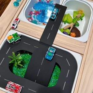 May include: A wooden sensory play table with toy cars on black road pieces, green grass, and colourful sensory bins filled with beads, glitter, and other objects. A fun and engaging toy for children.