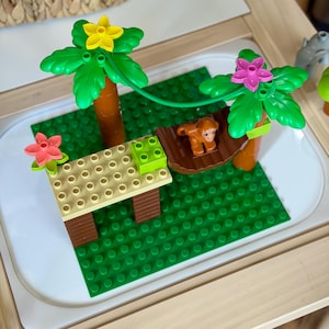 May include: A colorful jungle scene built with toy blocks. The scene includes palm trees with green fronds and flowers, a tan table, a brown monkey, and a green base. The blocks are arranged on a white tray.
