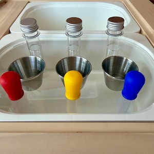 May include: A sensory play set featuring three clear test tubes with silver caps, three small stainless steel cups, and three colorful silicone stoppers in red, yellow, and blue. The set is arranged on a white surface within a wooden frame.