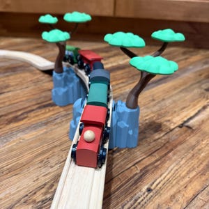 May include: A wooden toy train set with a red, green, and black engine and cars. The train is on a wooden track that curves around blue rock formations with green tree-shaped toppers. The set is on a wooden surface.