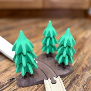 May include: Three green wooden toy trees on a brown base, with a wooden train track piece in the foreground. The trees have a detailed, layered design. The scene is set on a wooden surface.