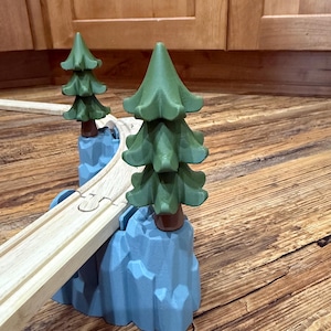 May include: Wooden toy train track with two green pine trees and blue mountain-shaped supports. The track is light brown, and the trees have a dark green color. The supports are a light blue color. The toy is designed for children.