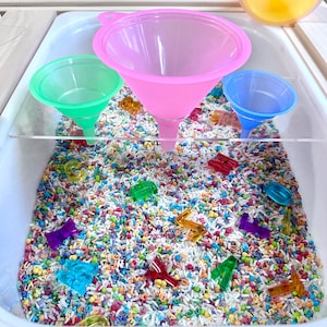 K&ouml;nnte beinhalten: Eine sensorische Wanne gef&uuml;llt mit buntem Reis und transparenten Kunststoffformen. Drei Trichter in Pink, Gr&uuml;n und Blau sind &uuml;ber dem Reis platziert. Die Wanne ist wei&szlig; und hat eine transparente Kunststoffabtrennung.