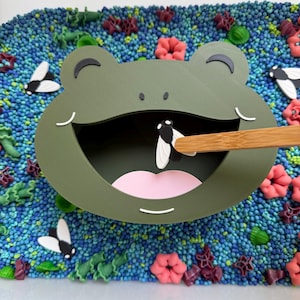 May include: A green frog-shaped toy with an open mouth, a pink tongue, and a fly being inserted with a wooden stick. The frog is surrounded by blue, green, and pink decorative elements, creating a playful scene.