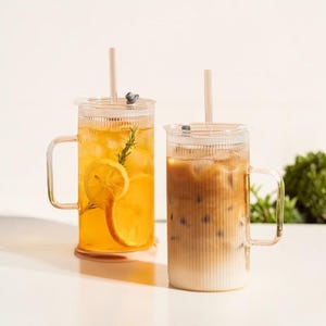 May include: Two clear glass mugs with lids and handles, filled with iced drinks. One contains a yellow drink with lemon slices and rosemary. The other has iced coffee. Both have straws, perfect for summer drinks.