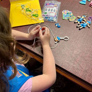 May include: A craft kit for making Girl Scout bead dolls, featuring a clear plastic bag with the text Girl Scout Beadolls, assorted colorful plastic beads, white cord, white lobster clasps, and several finished bead doll keychains.